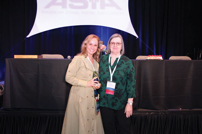 Two women smile for the camera. One wears a dress, and the other wears pants and a blouse. Both are wearing conference badges. Behind them is a stage with chairs and tables arranged for a speaker panel.