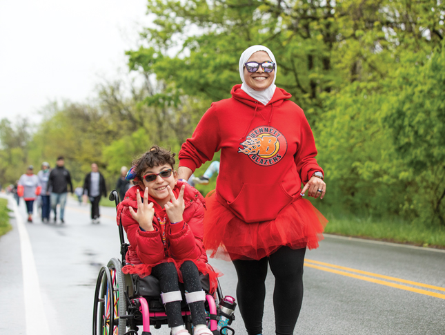 A smiling woman jogs on a road, pushing a smiling boy in a wheelchair. Both wear red hoodies that say “Bennett Blazers” on them, and both are wearing sunglasses.