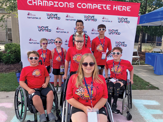 Posed photo of eight children and teens, some sitting in wheelchairs, and all wearing sunglasses and red T-shirts that say “Bennett Blazers.” Most are also wearing badges. They are posed in front of a banner that says “Move United Nationals” in multiple places.