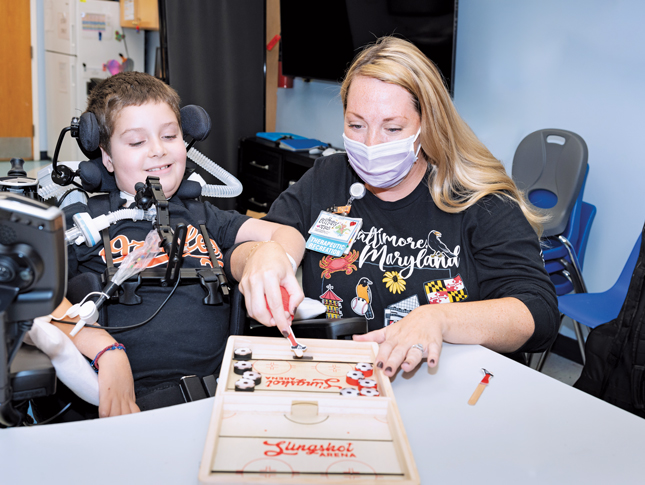 A boy is smiling as he works on an activity with a woman, who is wearing a face mask. The boy is sitting at a table in a wheelchair and has clear plastic tubes attached to his neck for breathing purposes. The woman is guiding the boy’s left hand through the activity.