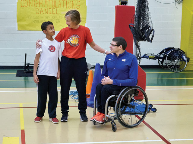 Gwena Herman, in a red Bennett Blazers T-shirt, stands with her right arm around the shoulders of a boy, Landon Brown, who wears a white Bennett Blazers T-shirt. Gwena’s left arm rests on the shoulder of a young man, Daniel Romanchuk, who sits in a wheelchair and wears a blue athletic zip-up shirt with a small USA flag on it.”