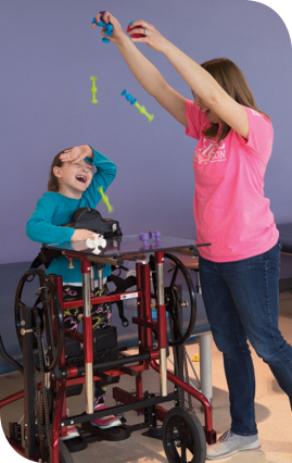 Physical therapist Brooke Meyer (right) drops suction cup toys on Caetlyn during physical therapy at Kennedy Krieger. A stander supports Caetlyn in an upright position.