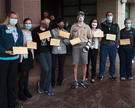 Eight people, all wearing face masks, stand in a line outside a Kennedy Krieger building, each one holding a wooden card holder. The fourth person from the right is a young man in a Boy Scout uniform.