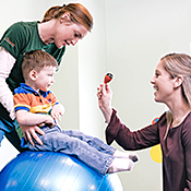 Physical therapist Kelly Fox helps Luke sit on a large blue therapy ball, while occupational therapist Catherine Syretz holds up an object for Luke to took at.