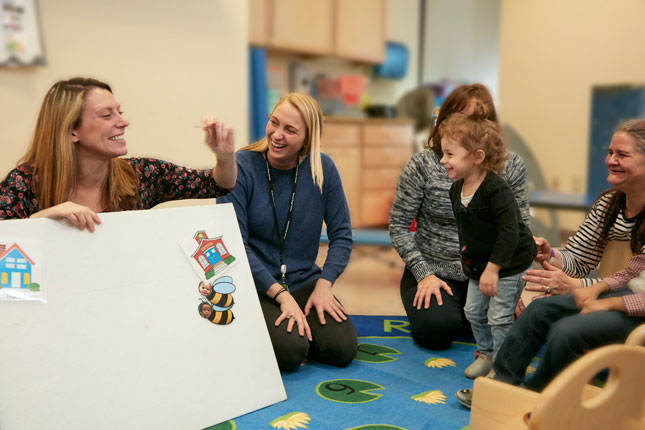 Speech Therapists sit on the floor and interact with a toddler and caretaker at the Child and Family Support Center.