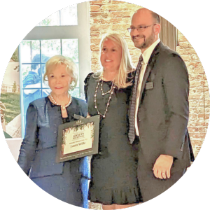Laura Wille receives the Mimi Baklor Volunteer of the Year Award. She is standing in the center, with a woman wearing a blue blazer and pearls to her left and a man in a suit to the right.