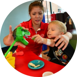 A young boy plays with a toy dinosaur as part of his therapy at Kennedy Krieger. His therapist sits his right, and she has her left arm placed on top of his left shoulder.