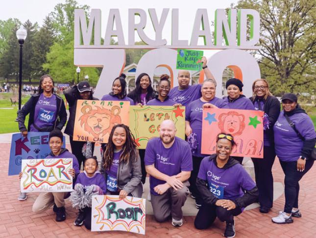 A group of BGE employees hold signs in support Kennedy Krieger's ROAR event at the Maryland Zoo. They are standing in front of a larger Maryland ZOO entrance smile.