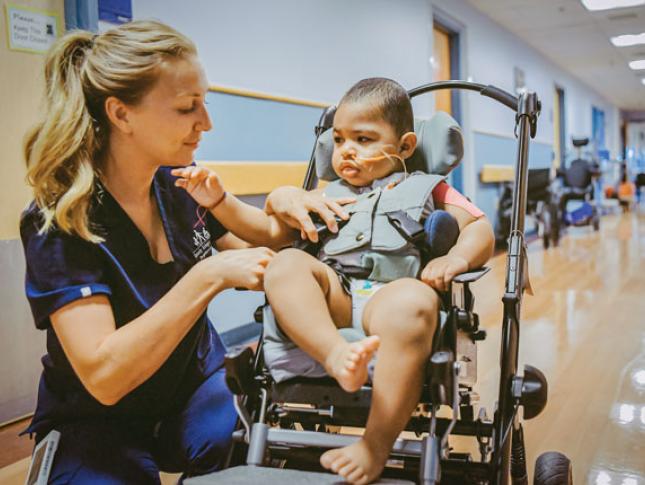 A nurse helps a young patient who is sitting in a mobility device.