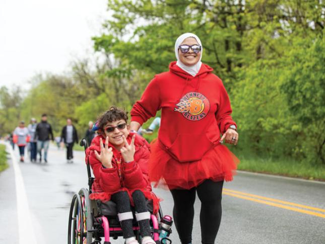 A child in a mobility device participates in a fun run and smiles. A woman is running along side them, and also smiling.