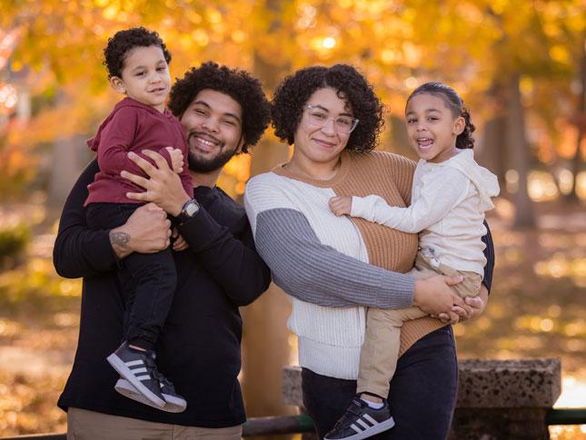 A family of four--father, son, mother an daughter--smile. The father is holding the son, while the mother is holding the daughter. They are standing outside with fall foliage visible in the background, 