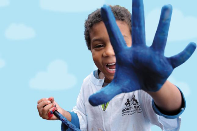 A child holds a paintbrush in his right hand while holding his left hand to the camera, with his palm covered in blue paint