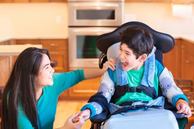 A young boy in a wheelchair shares a laugh with his teenage sister in their kitchen,