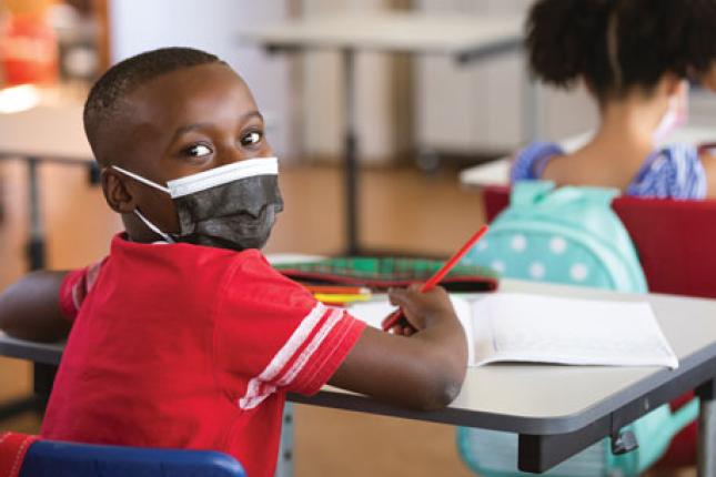 A young boy at his school desk looks over his shoulder at the camera while holding a pencil in his right hand and wearing a face mask
