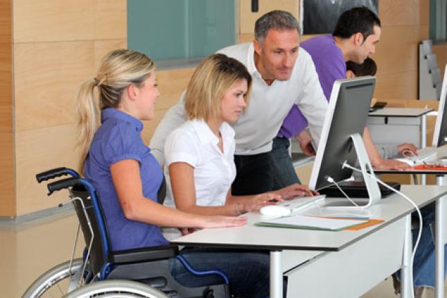 A woman in a wheelchair works at a shared desk. A woman sitting in a chair and a man standing over the desk are to her right, looking at a monitor. 