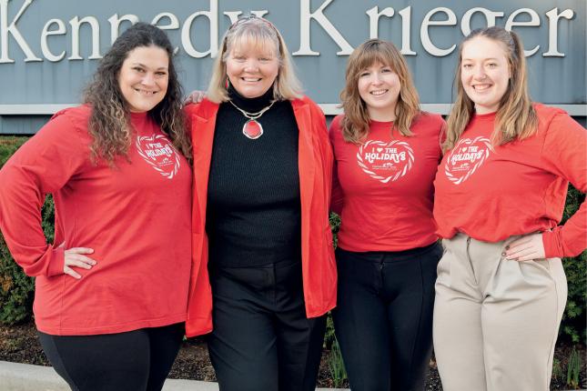 Four women stand outside in front of a sign that says “Kennedy Krieger.” Three of them are wearing red T-shirts that say “I love the holidays,” with the Kennedy Krieger Festival of Trees logo underneath.