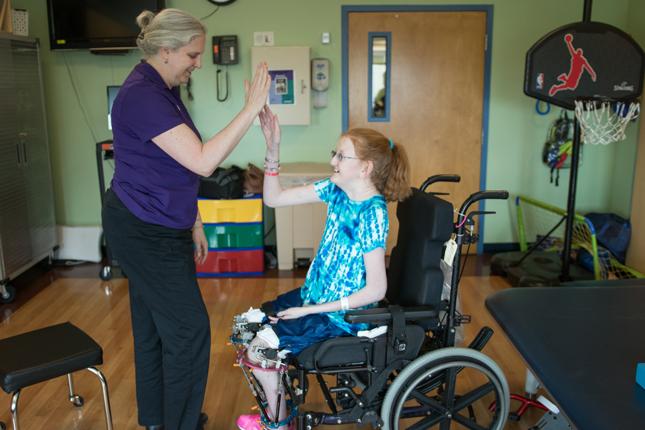 A teenaged girl in a blue and white shirt sits in her wheelchair and high fives her therapist, a woman wearing a purple shirt. 