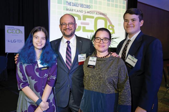 Family photo of a teenaged girl and boy with their parents. Father and son are each wearing a suit and tie, and mother and daughter are each wearing a dress. Each family member is wearing a name badge. Behind them is a screen that says “The Daily Record—Maryland’s Most Admired CEOs.”