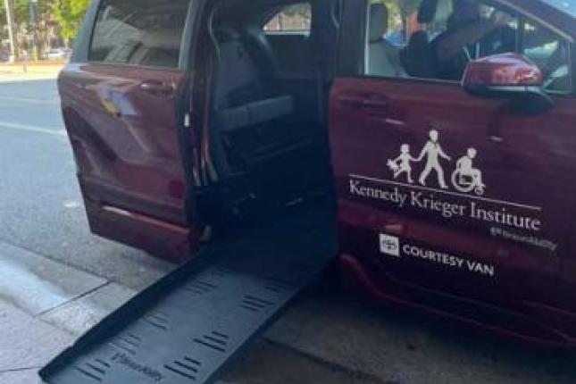 سيارة تويوتا سيينا يمكن الدخول إليها بواسطة كرسي متحرك مركونة عند الرصيف وتم إنزال الممر المنحدر. وتحمل السيارة المجهزة ذات اللون الأحمر الداكن شعار معهد Kennedy Krieger على باب الراكب الأمامي.