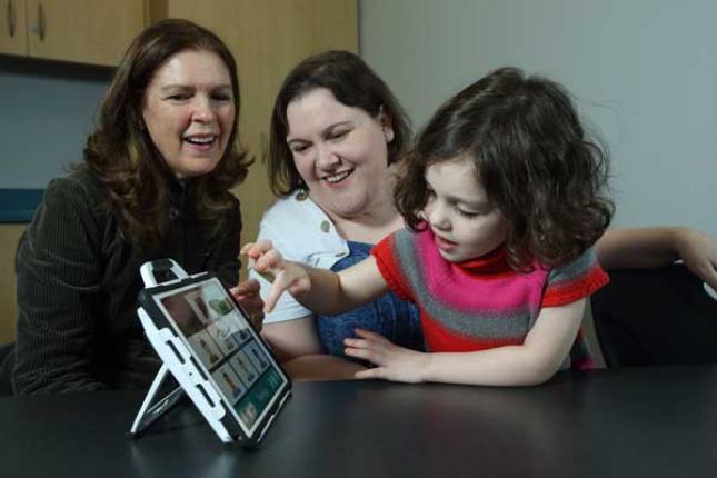 A young girl completes an activity using a tablet, while two women over her right shoulder smile.