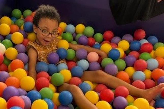 A young girl sits in a ball pit and smiles.