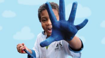 A child holds a paintbrush in his right hand while holding his left hand to the camera, with his palm covered in blue paint