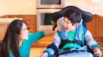 A young boy in a wheelchair shares a laugh with his teenage sister in their kitchen,