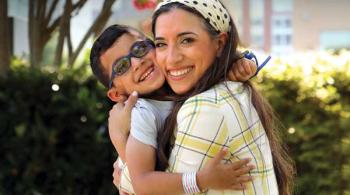 A young boy hugs his mom outside. Both have big smiles. The boy wears glasses and a kippa, and has a birthmark on his right ear and on the right side of his neck.