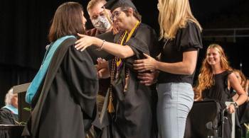 Hailey stands on the stage at her high school graduation and reaches out to hug her school principal. Hailey is smiling and wears a black graduation cap and gown, a medal, and purple and gold academic cords draped over her shoulders. Her school principal is also smiling and wears a black academic gown with a hood lined with light blue velvet. Two therapists stand to either side of Hailey, ready to help her if she needs assistance walking or standing. Behind the group is Hailey’s sister, who is smiling and h