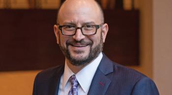 Professional headshot-style photo of a smiling white man with glasses and a beard, wearing a suit and purple tie.