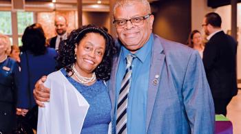 A woman and a man smile for the camera. The man is taller and to the right, and he has his right hand around the woman’s right shoulder. The woman is leaning into the man’s right shoulder. The man is wearing a blue suit, tie and glasses, and the woman is wearing a blue dress and pearl necklace.