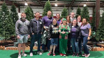 A group of 11 people of various ages, many wearing purple and one wearing a green Christmas elf costume, pose in front of a group of artificial trees inside a large indoor space, which looks like it might be a convention hall.