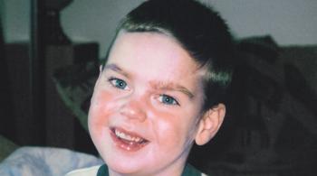 Un joven niño con ojos azul verdosos y cabello rubio sonríe para la foto. Lleva puesta una camiseta blanca con ribetes verdes oscuros.