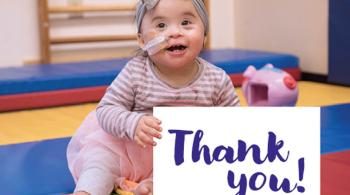 A toddler sits on the floor, with her left hand holding a white sign that says Thank you! in purple font.