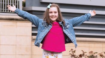 A young girl smiles with her arms outstretched. She has faint port wine-colored birthmarks across her forehead. A sign with the words “Kennedy Krieger” is in the background.