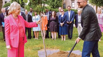 A woman and a man in business attire plant a tree, with a group of people standing behind them.