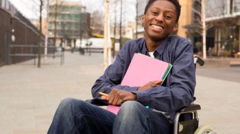 A young man smiles while sitting in a wheelchair and holding school supplies.