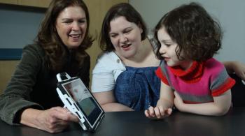 A doctor smiles while showing a little girl a game on a tablet. The girl is son her mother's lab, and the mother and doctor are seated at a table.