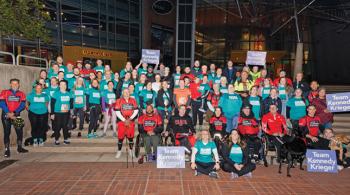 A large group of people, some wearing teal T-shirts and some wearing red T-shirts, pose for the camera. Some hold up purple signs that say “Team Kennedy Krieger.”