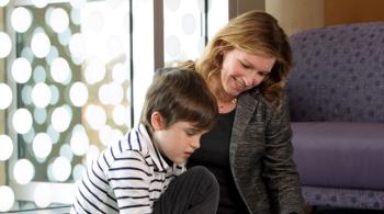 A boy with a muscular dystrophy sits on the floor next to a woman while reading a book.