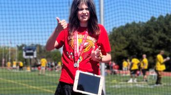 A girl wearing a augmentative and alternative communication device across her shirt stands up for a posed photo. She is participating in a Special Olympics event, and is also wearing a medal.