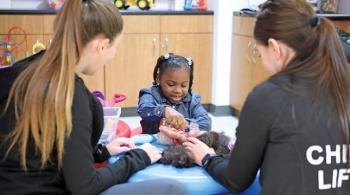Two child life specialists sit at a table and play a game with a little girl. The little girl is facing the camera, while both child life specialists have their backs to the camera.