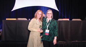 Two women smile for the camera. One wears a dress, and the other wears pants and a blouse. Both are wearing conference badges. Behind them is a stage with chairs and tables arranged for a speaker panel.